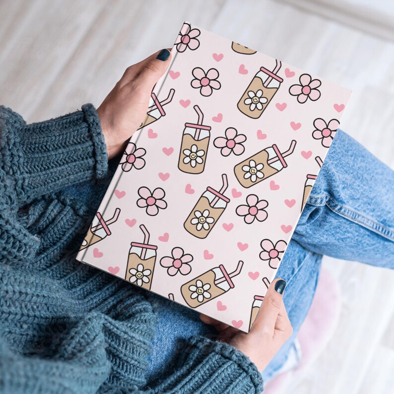 May include: A notebook with a pink cover featuring a repeating pattern of smiling iced coffee drinks, pink flowers, and hearts. The notebook is held open, revealing the patterned design. The overall theme is a cute, whimsical design.