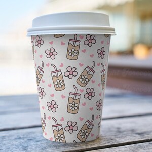 May include: A white paper coffee cup with a white lid. The cup is decorated with a repeating pattern of pink flowers, pink hearts, and brown iced coffee drinks with smiling flower faces.