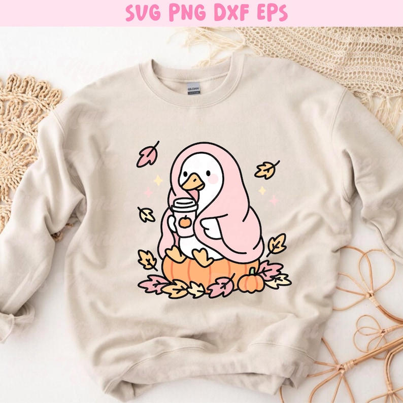 May include: Beige sweatshirt featuring a cartoon duck wrapped in a pink blanket, holding a coffee cup with a pumpkin design. The duck is sitting on a pumpkin surrounded by autumn leaves and small pumpkins. The text "SVG PNG DXF EPS" is at the top.