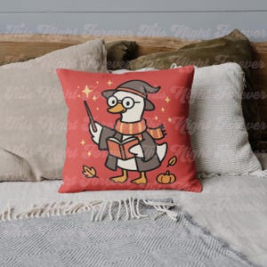 May include: Red decorative pillow featuring a cartoon duck dressed as a wizard, wearing glasses, a hat, and a scarf. The duck is holding a wand and a book. The pillow has a fall theme with leaves and a pumpkin.