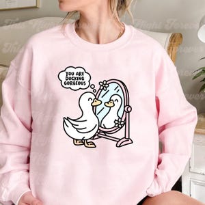 K&ouml;nnte beinhalten: Rosafarbener Pullover mit einer wei&szlig;en Ente, die in einen Spiegel schaut, mit dem Text "You are ducking gorgeous" in einer Gedankenblase &uuml;ber der Ente.
