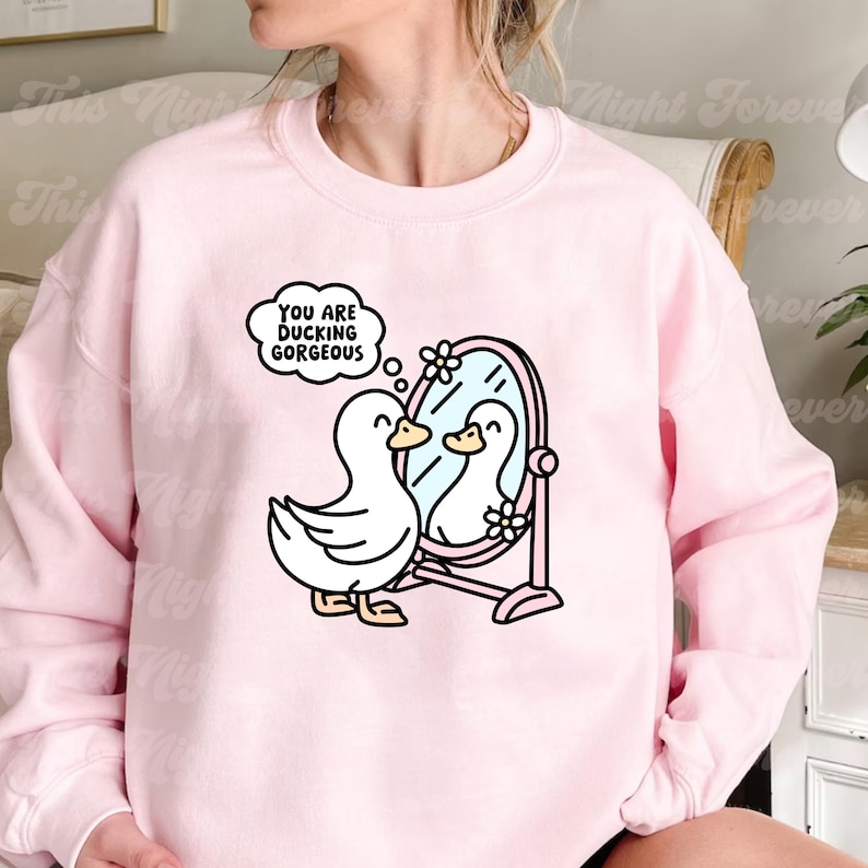 K&ouml;nnte beinhalten: Rosafarbener Pullover mit einer wei&szlig;en Ente, die in einen Spiegel schaut, mit dem Text "You are ducking gorgeous" in einer Gedankenblase &uuml;ber der Ente.