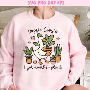 以下が含まれることがあります： 「Oopsie Goosie I got another plant」の文字と、植木鉢を持ったガチョウのグラフィックが描かれた、薄いピンク色のスウェットシャツ。デザインには、いくつかの鉢植えと装飾的な要素が含まれています。
