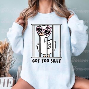 May include: A light blue sweatshirt with a cartoon illustration of a white goose wearing pink heart-shaped sunglasses and a pink bow. The goose is in a cage with the text "GOT TOO SILLY" below it.