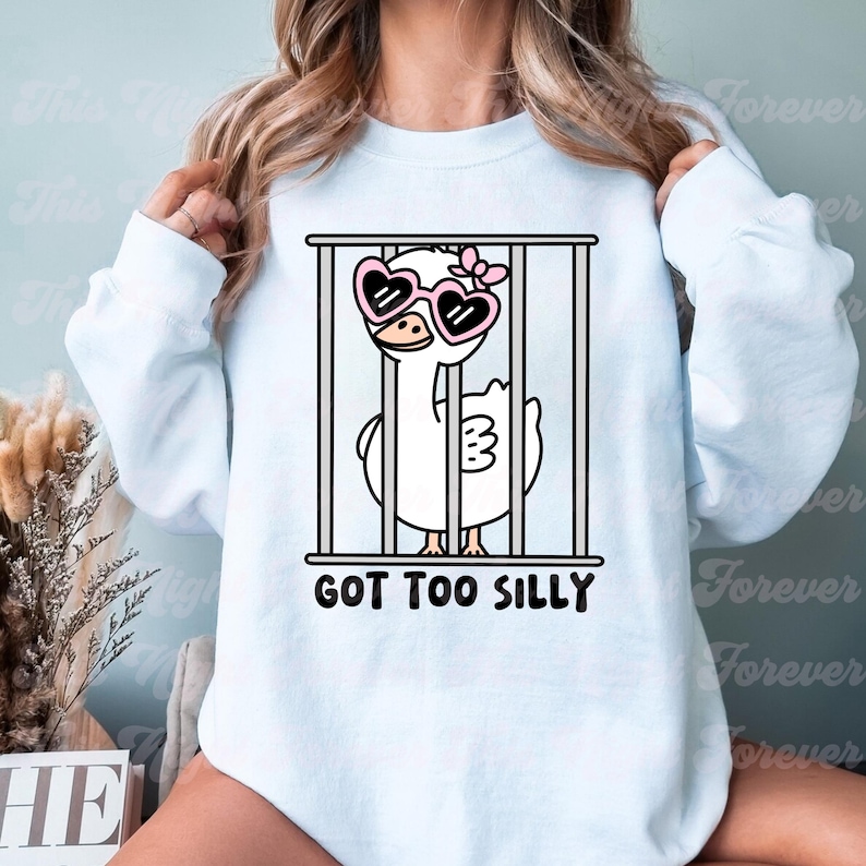 May include: A light blue sweatshirt with a cartoon illustration of a white goose wearing pink heart-shaped sunglasses and a pink bow. The goose is in a cage with the text "GOT TOO SILLY" below it.