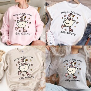 K&ouml;nnte beinhalten: Verschiedene Sweatshirts und ein wei&szlig;es T-Shirt mit einer Cartoon-Gans auf Rollschuhen. Die Gans hat eine rosa Schleife und ist von Blumen- und Sterngrafiken umgeben. Der Text "sorry I'm late I was dilly dallying" ist aufgedruckt.