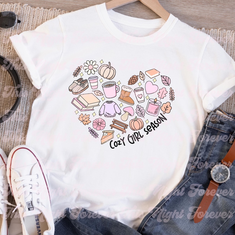 Op de afbeelding: Wit T-shirt met een roze hartontwerp met verschillende herfstthema-illustraties, waaronder een pompoen, bladeren, een mok koffie en een boek. De tekst "Cozy Girl Season" is geschreven in een roze handschrift.