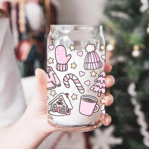 May include: A clear glass can-shaped cup decorated with pink winter-themed illustrations. The design includes a mitten, a beanie, a house, a candy cane, a tree, a mug, hearts, and stars. The cup is held in front of a blurred background.