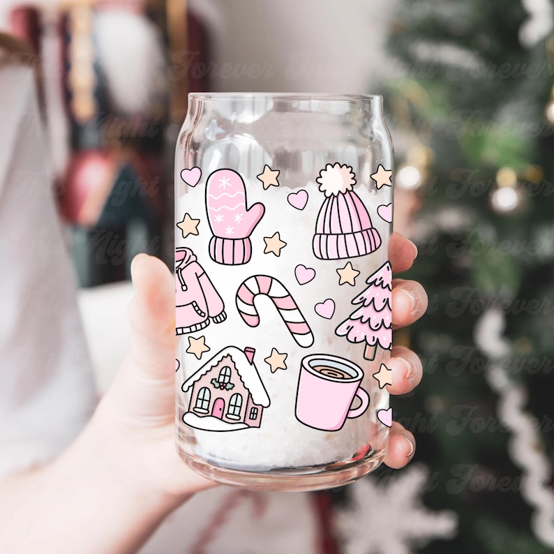 May include: A clear glass can-shaped cup decorated with pink winter-themed illustrations. The design includes a mitten, a beanie, a house, a candy cane, a tree, a mug, hearts, and stars. The cup is held in front of a blurred background.