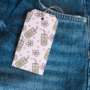 May include: A white gift tag with a repeating pattern of pink hearts, flowers, and iced coffee drinks. The tag is made of paper and has a hole at the top with a string attached. The tag is set against a blue denim background.
