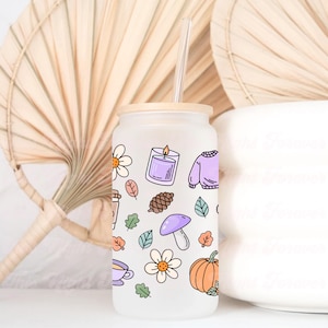 May include: Frosted glass tumbler with a bamboo lid and a clear straw. The tumbler is decorated with fall-themed illustrations, including a candle, sweater, pumpkin, and leaves. The design features a variety of colors, including orange, purple, and green.
