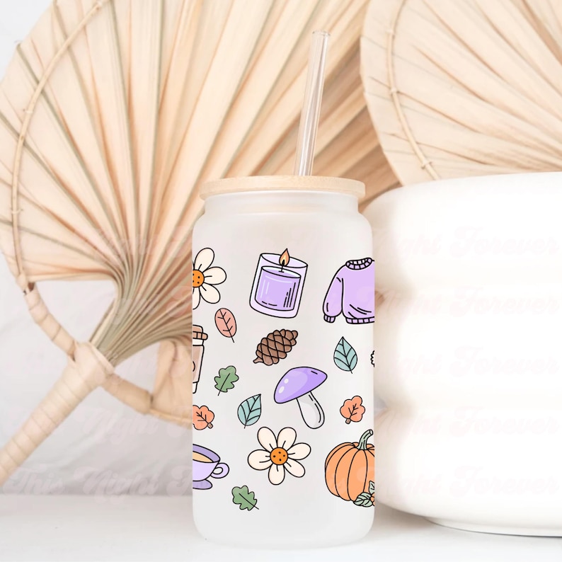 May include: Frosted glass tumbler with a bamboo lid and a clear straw. The tumbler is decorated with fall-themed illustrations, including a candle, sweater, pumpkin, and leaves. The design features a variety of colors, including orange, purple, and green.