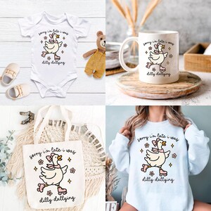 K&ouml;nnte beinhalten: Eine Auswahl an Artikeln mit einer Cartoon-Ente auf Rollschuhen und dem Text "sorry I'm late I was dilly dallying." Zu den Artikeln geh&ouml;ren ein wei&szlig;er Baby-Body, eine wei&szlig;e Tasse, eine wei&szlig;e Tragetasche und ein hellblaues Langarmshirt.