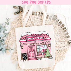 May include: A white tote bag featuring a pink illustration of a bookstore with the text "BOOK STORE" on the sign. The design includes a Christmas tree, snow, and the words "cozy vibes" on a small chalkboard.