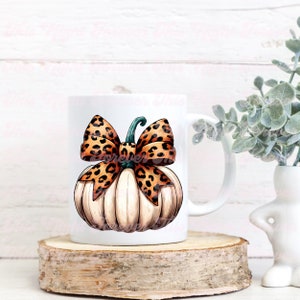 May include: White ceramic mug with a hand-painted design of a white pumpkin with a brown and orange leopard print bow. The pumpkin has a green stem.