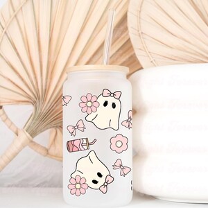 May include: A frosted glass tumbler with a bamboo lid and a clear straw. The tumbler is decorated with pink bows, flowers, and cartoon ghosts. A pink drink illustration is also featured on the glass. The tumbler is a cute and whimsical design.