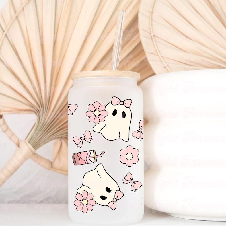 May include: A frosted glass tumbler with a bamboo lid and a clear straw. The tumbler is decorated with pink bows, flowers, and cartoon ghosts. A pink drink illustration is also featured on the glass. The tumbler is a cute and whimsical design.
