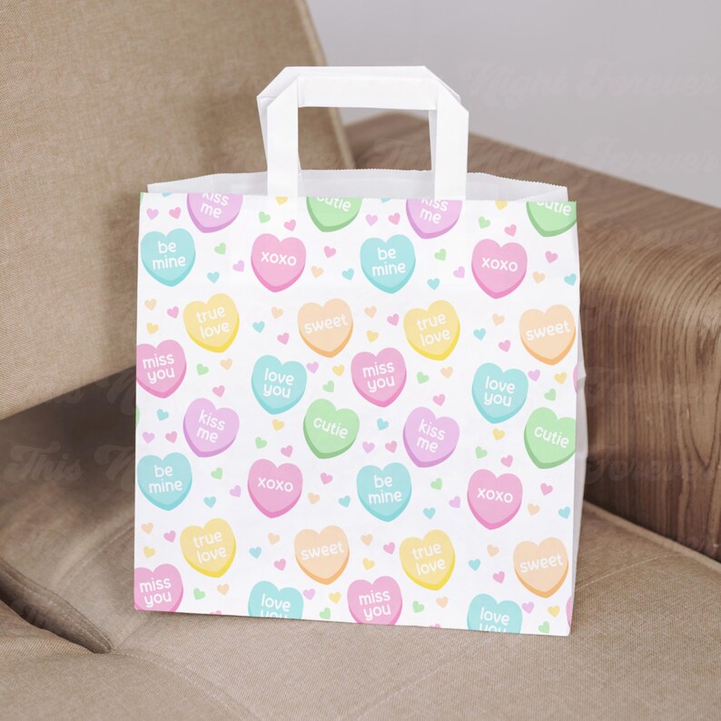 May include: White paper gift bag with a colorful heart pattern. The hearts have various messages, including "kiss me", "true love", "be mine", "miss you", "love you", "cutie", "sweet", and "xoxo".
