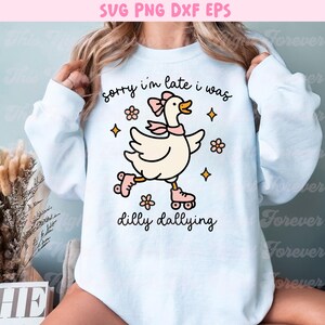 K&ouml;nnte beinhalten: Wei&szlig;es Sweatshirt mit einer Cartoon-Gans auf Rollschuhen, mit rosa Schleife und Schal. Der Text "sorry i'm late i was dilly dallying" ist in schwarzer Schrift geschrieben. Blumen- und Sterngrafiken umgeben die Gans.