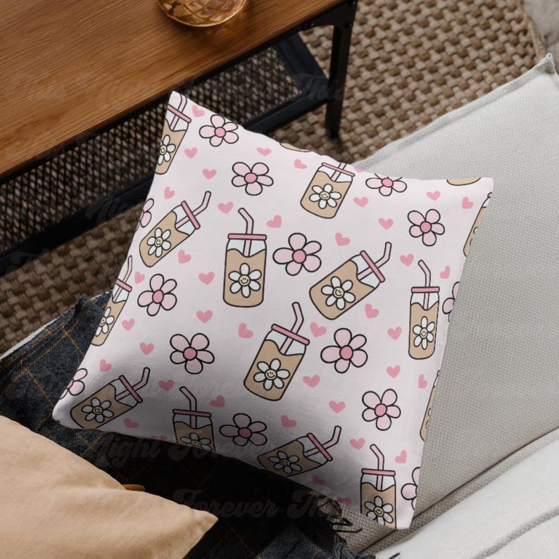 May include: Square throw pillow with a repeating pattern of iced coffee drinks, flowers, and hearts in shades of beige, pink, and white. The pillow is on a light-colored couch.