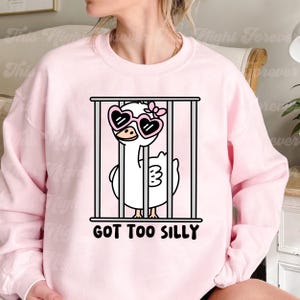 May include: Pink sweatshirt with a white cartoon goose wearing heart-shaped sunglasses and a bow. The goose is in a cage with the text "GOT TOO SILLY".
