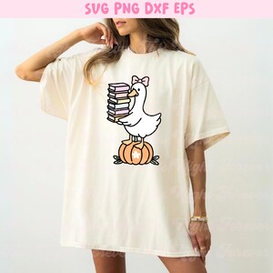 May include: Cream-colored t-shirt featuring a cartoon duck with a pink bow holding a stack of books. The duck is standing on an orange pumpkin with green leaves. The text "SVG PNG DXF EPS" is at the top.