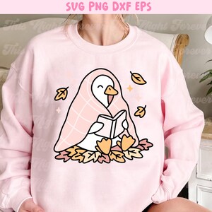 May include: A light pink sweatshirt featuring a cartoon goose reading a book while wrapped in a pink blanket. The goose is surrounded by autumn leaves and falling leaves. The top of the image has the text "SVG PNG DXF EPS".