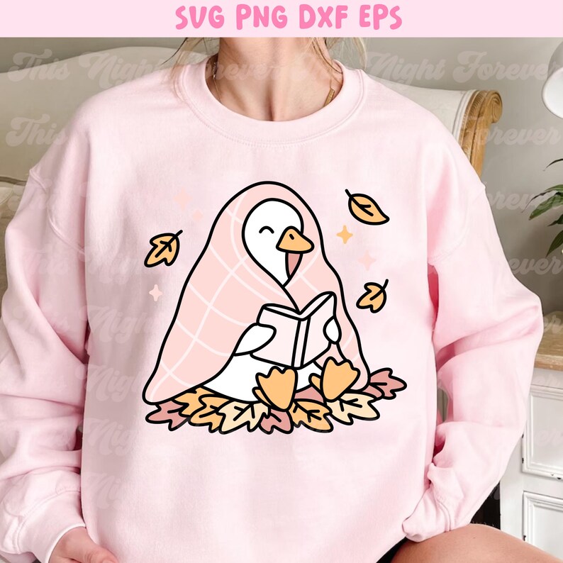 May include: A light pink sweatshirt featuring a cartoon goose reading a book while wrapped in a pink blanket. The goose is surrounded by autumn leaves and falling leaves. The top of the image has the text "SVG PNG DXF EPS".