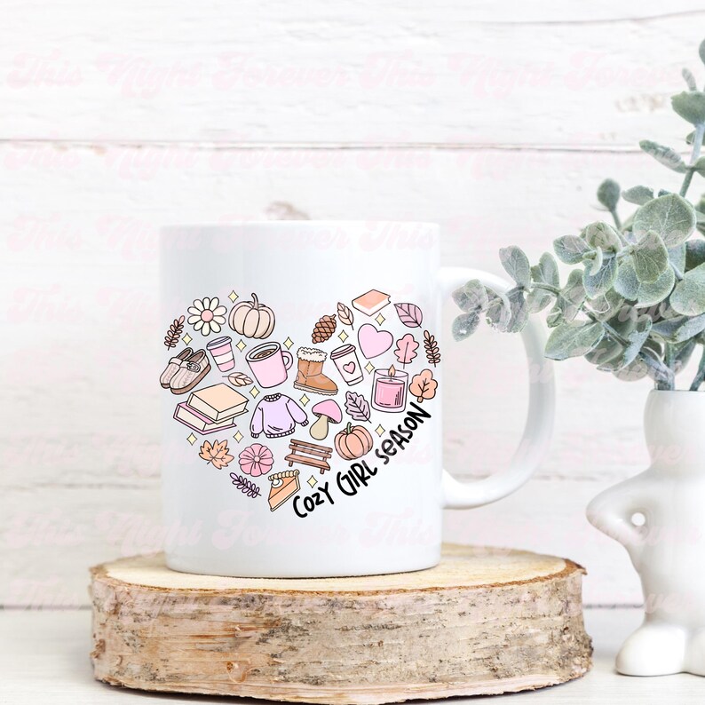Op de afbeelding: Witte keramische mok met een hartvormig ontwerp met verschillende herfstthema-illustraties, waaronder bladeren, pompoenen, laarzen en een boek. De tekst "Cozy Girl Season" is geschreven in een roze handschrift.