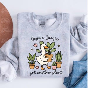 以下が含まれることがあります： 「Oopsie Goosie I got another plant」の文字が入ったライトグレーのスウェットシャツ。漫画のガチョウが植木鉢を持っており、他の鉢植えの植物や花のアクセントに囲まれています。