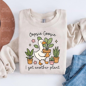以下が含まれることがあります： ベージュ色のスウェットシャツ。アヒルが鉢植えの植物を持っているイラストが描かれています。「Oopsie Goosie I got another plant」の文字がアヒルの上下にプリントされています。他の鉢植えの植物や花も描かれています。