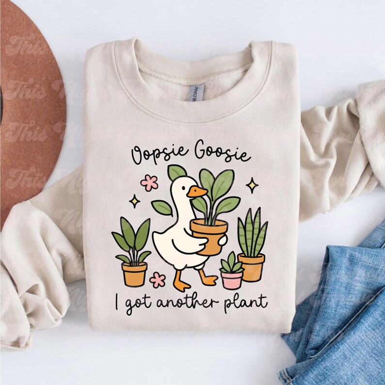 以下が含まれることがあります： ベージュ色のスウェットシャツ。アヒルが鉢植えの植物を持っているイラストが描かれています。「Oopsie Goosie I got another plant」の文字がアヒルの上下にプリントされています。他の鉢植えの植物や花も描かれています。