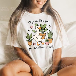 以下が含まれることがあります： 白いTシャツ。漫画のガチョウが鉢植えの植物を持っており、他の鉢植えの植物と「Oopsie Goosie I got another plant」というテキストに囲まれています。デザインにはピンクの花と星が含まれています。