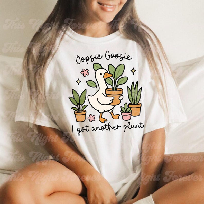 以下が含まれることがあります： 白いTシャツ。漫画のガチョウが鉢植えの植物を持っており、他の鉢植えの植物と「Oopsie Goosie I got another plant」というテキストに囲まれています。デザインにはピンクの花と星が含まれています。