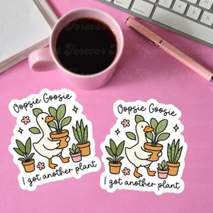 以下が含まれることがあります： 「Oopsie Goosie」と「I got another plant」の文字が入った2つの白いビニールステッカー。各ステッカーには、鉢植えの植物を運ぶ漫画のガチョウが描かれており、他の鉢植えの植物や花のアクセントに囲まれています。ピンクのマグカップとペンも表示されています。