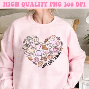 Op de afbeelding: Roze sweatshirt met een hartvormig ontwerp met verschillende herfstthema-illustraties, waaronder een pompoen, bladeren, een mok koffie, een boek en een taart. De tekst "Cozy Girl Season" staat in het hart.