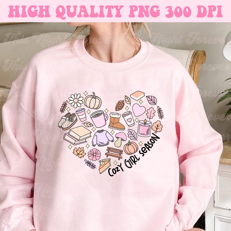Op de afbeelding: Roze sweatshirt met een hartvormig ontwerp met verschillende herfstthema-illustraties, waaronder een pompoen, bladeren, een mok koffie, een boek en een taart. De tekst "Cozy Girl Season" staat in het hart.