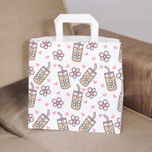 May include: White paper gift bag with a repeating pattern of cartoon iced coffee drinks, pink flowers, and hearts. The bag has a white handle and is sitting on a neutral-colored surface.