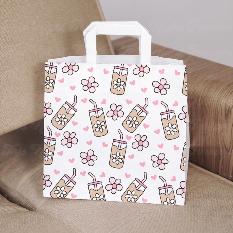May include: White paper gift bag with a repeating pattern of cartoon iced coffee drinks, pink flowers, and hearts. The bag has a white handle and is sitting on a neutral-colored surface.