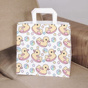 May include: White paper gift bag with a repeating pattern of yellow ducks, pink inner tubes, pastel-colored beach balls, and stars. The bag has a white handle and is suitable for gifts.