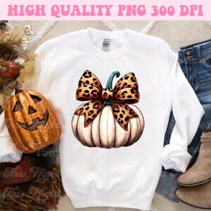 May include: White pumpkin with a large orange and brown leopard print bow. The pumpkin is sitting on a white sweatshirt.