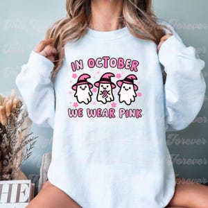 May include: A light blue sweatshirt with the text "In October We Wear Pink" and a graphic of three ghosts wearing pink witch hats and pink ribbons.