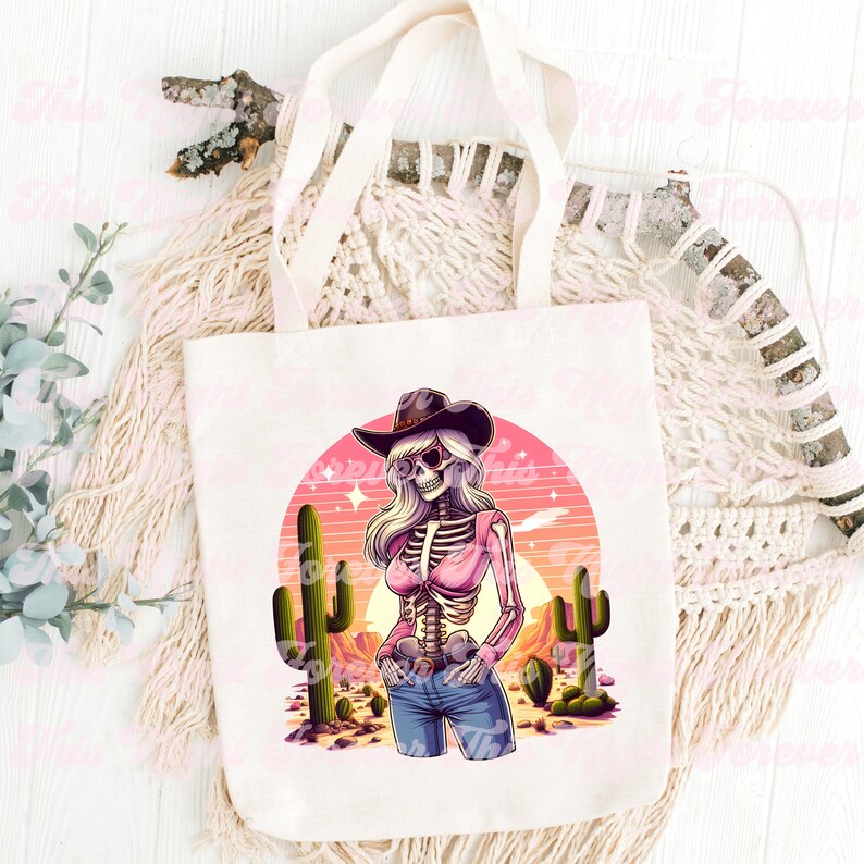 May include: Off-white canvas tote bag featuring a colorful illustration of a skeleton cowgirl wearing a hat and sunglasses, set against a desert sunset with cacti. The design includes a pink top and blue jeans.