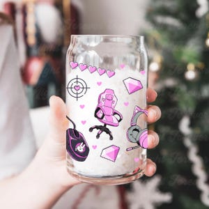May include: A clear glass can-shaped cup with pink gaming-themed graphics. The design includes a pink gaming chair, a mouse, a headset, hearts, and a target symbol. The cup is held in a person's hand.