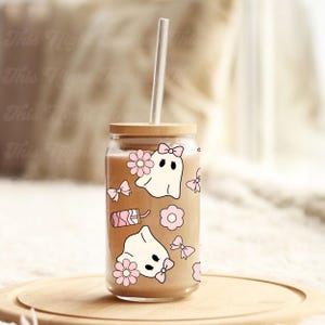 May include: A clear glass tumbler with a wooden lid and a clear straw. The tumbler is decorated with pink bows, flowers, and ghost illustrations. The glass contains a light brown liquid.