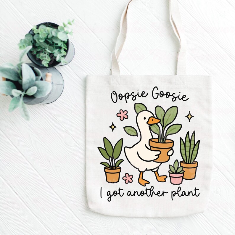 以下が含まれることがあります： クリーム色のトートバッグ。アヒルが植木鉢を持ったイラストが描かれています。「Oopsie Goosie」と「I got another plant」の文字がプリントされています。アヒルの周りには、植木鉢に入った植物や花の装飾が施されています。