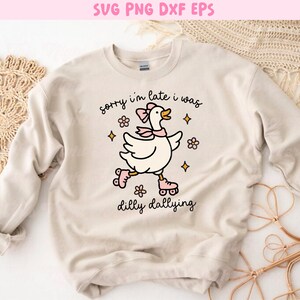 K&ouml;nnte beinhalten: Cremefarbenes Sweatshirt mit einer Cartoon-Gans auf rosa Rollschuhen, mit Schleife und Schal. Der Text lautet "sorry i'm late i was dilly dallying". Das Design umfasst Blumen und Sterne.