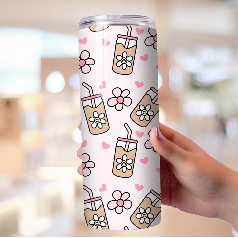 May include: A white tumbler with a repeating pattern of cartoon iced coffee drinks with smiling flower faces, pink flowers, and pink hearts. The tumbler has a clear lid and a straw. The design is on a light pink background.