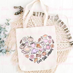 Op de afbeelding: Een witte canvas tote bag met een roze en bruin hartontwerp met herfstthema illustraties. De tekst "Cozy Girl Season" staat in zwart in het hart.