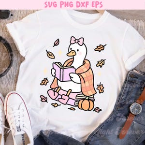 May include: White t-shirt featuring a cartoon goose reading a book, wrapped in a plaid scarf, and surrounded by autumn leaves. The goose wears a pink bow and is sitting on books with a pumpkin. Text at the top reads "SVG PNG DXF EPS".